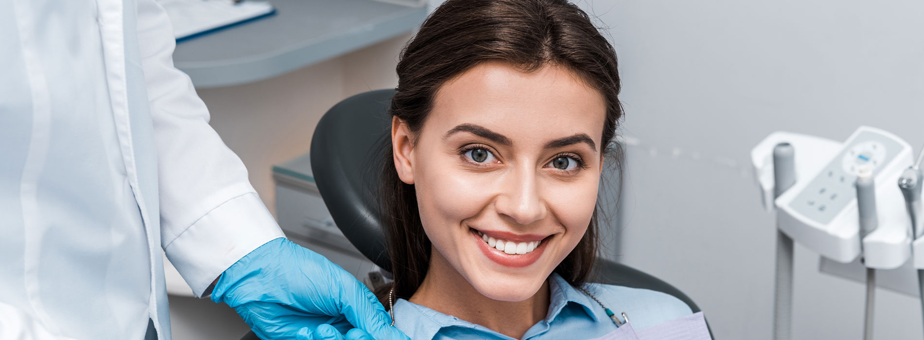 The image shows a woman with a bright smile sitting in a dental chair, receiving dental care from a professional wearing protective gear, including gloves and a face mask.