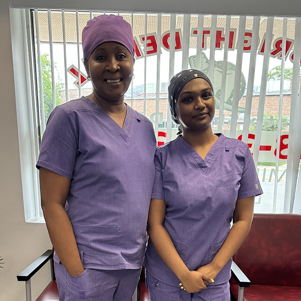 Two women wearing scrubs are standing side by side in front of a sign with the word hospital on it, posing for a photo.