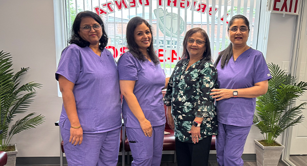 The image shows five individuals posing together in an office setting they are dressed in scrubs, suggesting they may be healthcare professionals, standing in front of a desk with a chair and a potted plant, with a sign that reads Welcome visible behind them.