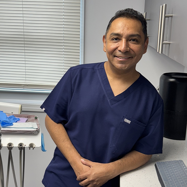 The image shows a man wearing scrubs, standing with his hands together in front of him, smiling at the camera. He appears to be in a clinical setting, possibly an office or exam room, as suggested by the presence of dental equipment and a chair with what looks like a blue cloth draped over it.