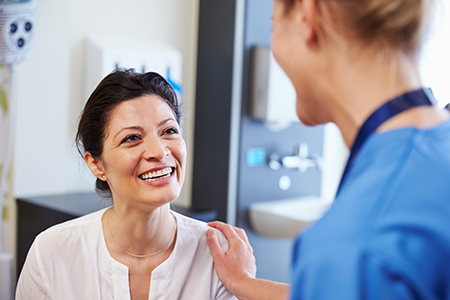 The image shows a woman with dark hair, wearing a white shirt, smiling at a male healthcare professional who appears to be a dentist, standing in an office setting.