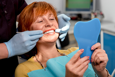 The image shows a woman sitting in a dental chair with her mouth open, holding up a blue model of teeth for comparison, while a dentist attends to her with tools and wearing gloves.