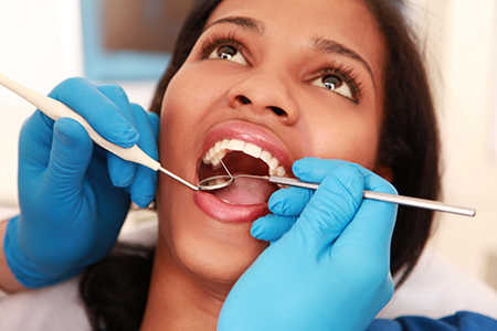 The image shows a person receiving dental care, with a dental professional performing work on their teeth while wearing blue gloves and protective eyewear.