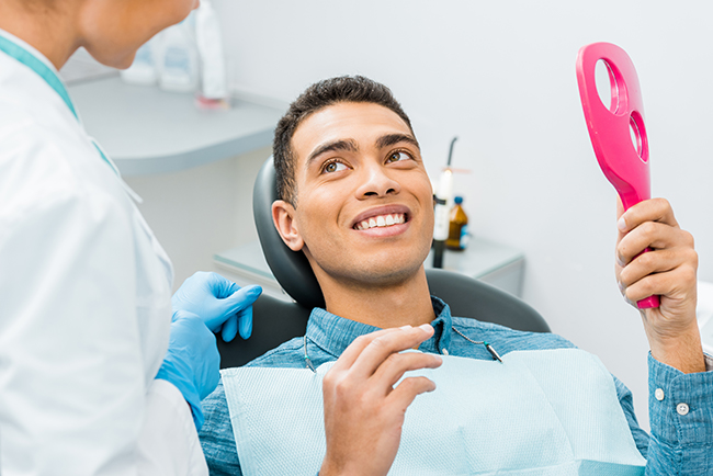 The image shows a smiling man seated in a dental chair, receiving dental care from a professional who is holding a pink electric toothbrush.