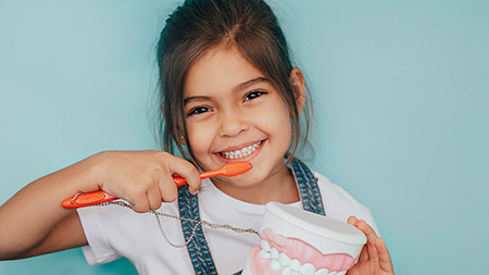 The image shows a young girl with a big smile brushing her teeth while holding a toothbrush in her hand, with two separate images side by side.