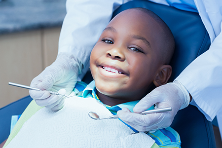 A young boy sitting in a dental chair with a smile, receiving dental treatment from a dentist wearing gloves and a blue apron.