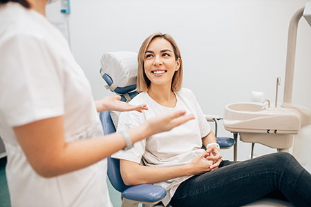 The image shows a woman sitting in a dental chair, smiling at the camera, with a dental professional standing beside her, both in a dental office setting.